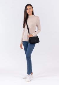 Young woman standing wearing beige long-sleeve top, blue jeans, white sneakers, and carrying a black crossbody bag.