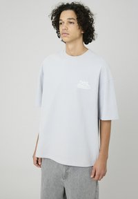 T-shirt en coton gris clair à manches courtes et col rond, doté d'un graphique blanc "Rayonner la Positivité" sur la zone gauche de la poitrine.