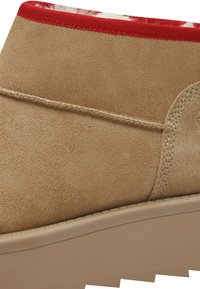 Beige-nahkaiset saappaat, joissa on teksturoitu pinta, punaisella reunalla ja vaalealla pohjalla. Sisältää tikkausyksityiskohtia saumoissa.