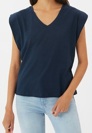 Frau trägt ein ärmelloses, dunkelblaues V-Ausschnitt-Oberteil und hellblaue Jeans, steht vor einem schlichten weißen Hintergrund.