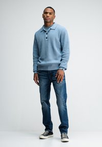 Helleblaue gerippte Strickjacke mit kragenförmigem Ausschnitt und drei Knöpfen, kombiniert mit Jeans in mittlerer Waschung und schwarzen Turnschuhen.