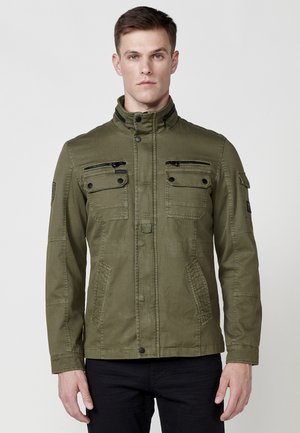 Chaqueta verde oliva, con un cuello alto, múltiples bolsillos con cremallera y de solapa, y una tela texturizada. Diseño minimalista sin patrones visibles.