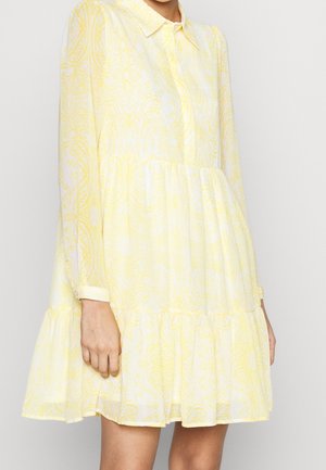 Femme portant une robe courte à manches longues jaune et blanche, à motifs, avec un col et un ourlet à volants.