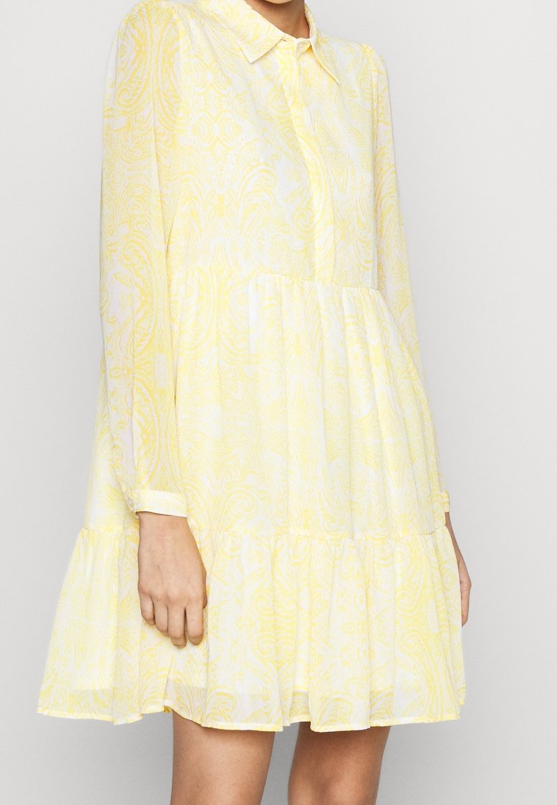 Femme portant une robe courte à manches longues jaune et blanche, à motifs, avec un col et un ourlet à volants.