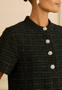 Czarna teksturowana kurtka z krótkimi rękawami, z okrągłym kołnierzem i trzema dekoracyjnymi guzikami w kształcie kwiatów z kryształowymi detalami.
