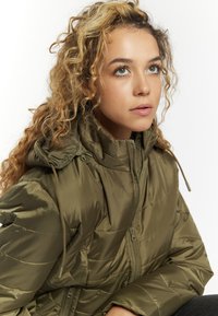 Giacca imbottita verde oliva con cappuccio, dotata di zip frontale, texture trapuntata e coulisse elastiche sul cappuccio e in vita.