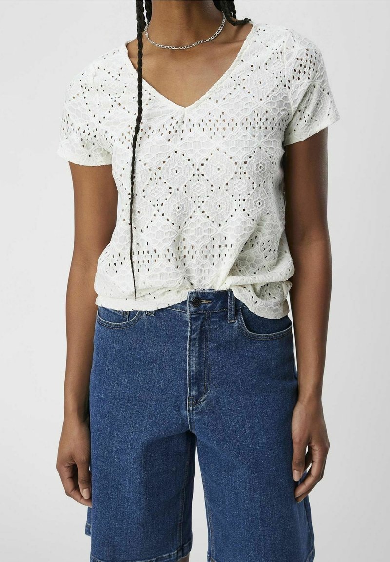 Weißes kurzärmliges Oberteil mit V-Ausschnitt, mit einem floralen Lochmuster. Kombiniert mit dunkelblauen Denim-Shorts, die eine lockere Passform und Taschen haben.