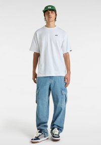 Weißes T-Shirt mit kurzen Ärmeln und kleinem Logo, hellblaue Cargo-Hose mit Taschen und grüner Kappe; Marineblaue und weiße Sneakers.