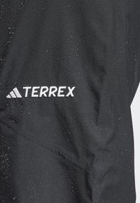 Veste noire imperméable avec des gouttes d'eau à la surface, arborant le logo "TERREX" en blanc. Le tissu semble texturé et durable.