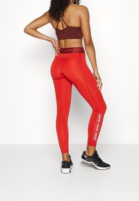 Červené legíny s úzkým střihem a černým páskem s logem "NIKE", doplněné hnědým sportovním podprsenkou a černými teniskami.