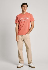 Korálová bavlnená tričko s potlačou "PEPE JEANS LONDON W11" bielou farbou, spárované s béžovými nohavicami a bielymi teniskami s hnedými podrážkami.