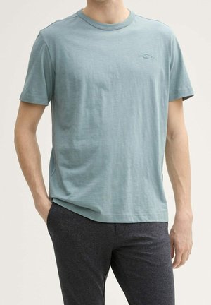 Lichtblauw T-shirt met korte mouwen van zacht materiaal heeft een ronde halslijn en een klein logo op de voorkant, gecombineerd met donkere gestructureerde broeken.