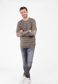 Uomo sorridente con tatuaggi su entrambe le braccia incrociate, indossa un maglione a maniche corte marrone, jeans grigi usurati e sneaker bianche.