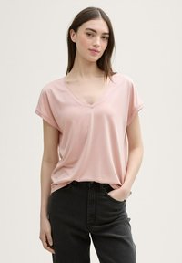 Tricou cu decolteu în V, mânecă scurtă, de culoare roz pal, fabricat dintr-un material moale, asortat cu blugi negri. Tricoul se așează lejer, având o croială relaxată.