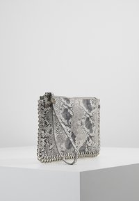 Malá obdélníková kabelka na ruku se šedým a černým hadím vzorem, kovovým řetízkovým lemem a zápěstním popruhem na bílé podstavě na šedém pozadí.