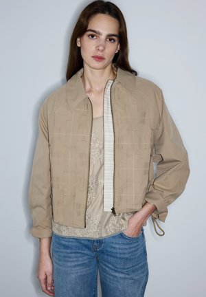 Młoda kobieta z długimi brązowymi włosami, ubrana w beżową plecioną kurtkę na floralnym topie i niebieskich dżinsach, stoi z jedną ręką w kieszeni.