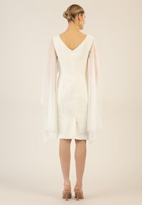 White dress with a fitted silhouette, V-back, and sheer, flowing sleeves. The knee-length design features a back zip and slight slit.