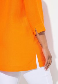 Felle oranje linnen top met driekwartmouwen met inkepingen, een rechte zoom en een losse pasvorm, gecombineerd met witte broeken.