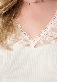 Witte ribgebreide top met een V-hals en kantafwerking. De zachte textuur en delicate kantaccenten voegen detail toe aan de halslijn.