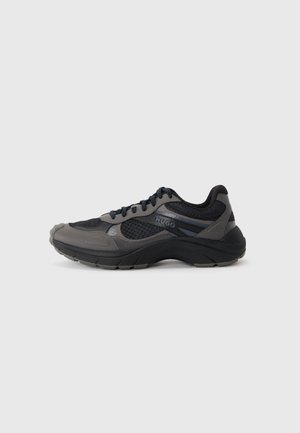 Sneaker HUGO nera e grigia con tomaia in mesh e pelle, lacci neri e suola nera spessa mostrata di profilo su sfondo grigio chiaro.
