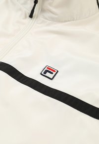 Chaqueta cortavientos blanca con cremallera en la parte delantera, franja horizontal negra y logotipo bordado en rojo y azul. Textura sintética suave.