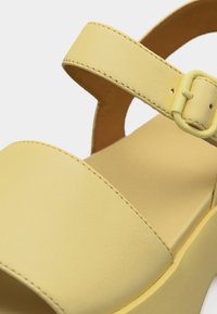 Κίτρινο σανδάλι πλατφόρμα με φαρδιά μπροστινή λωρίδα και ρυθμιζόμενο λουράκι αστραγάλου με πόρπη, κατασκευασμένο από λείο υλικό που μοιάζει με δέρμα.