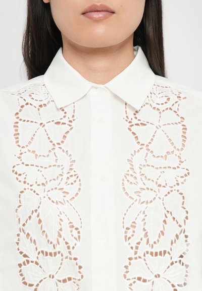 Camisa blanca con intrincados bordados de flores recortados en la parte frontal, con cuello clásico y cierre de botones. Textura de tela suave.
