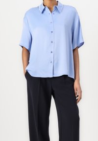 Světle modrá halenka s krátkým rukávem, zapínáním na knoflíky vpředu, límečkem a volnějším střihem. Hladká textura s jemným leskem. Nošena s tmavými kalhotami.