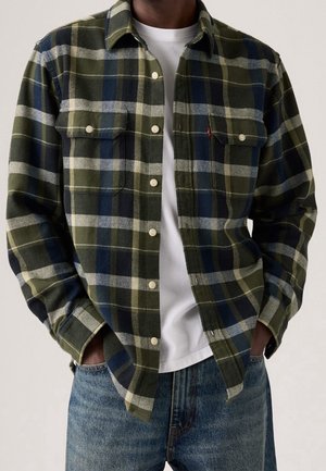 Homme portant une chemise à carreaux verte, bleu marine et blanche par-dessus un T-shirt blanc avec un jean bleu, mains dans les poches.