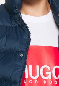 Námořnická prošívaná bunda s vysokým límcem, zipovým zapínáním a patentkami. Nositá přes bílý tričko s červeným logem. Hladký povrch.