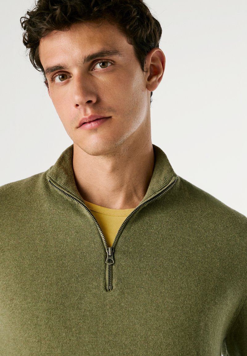 Zielona sweter z zamkiem, wykonany z miękkiego materiału, z kołnierzem stojącym. Nosi się go na żółtej koszulce, o delikatnej teksturze i bezszwowym wzorze.