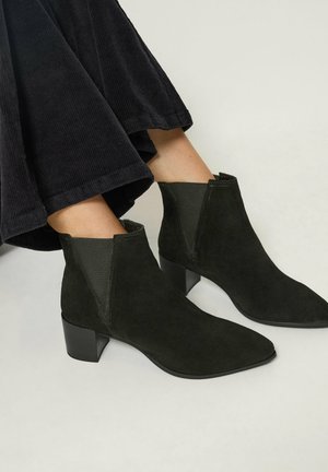 Pieds portant des bottines en daim noir à talons blocs bas et un pantalon noir à jambes larges sur un fond uni.