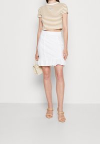 Pruhované béžové zkrácené tričko s trojúhelníkovým logem, spárované s bílou sukní z krajky s volánkovým lemem. Béžové sandály na řemínky a kabelka.