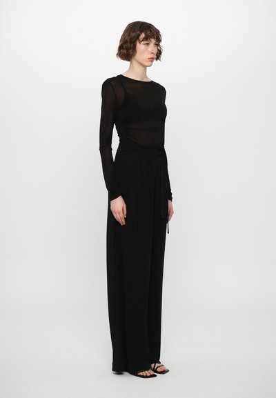 Black long-sleeve top in sheer fabric paired with high-waisted black trousers, featuring a tied waist detail and flat sandals.