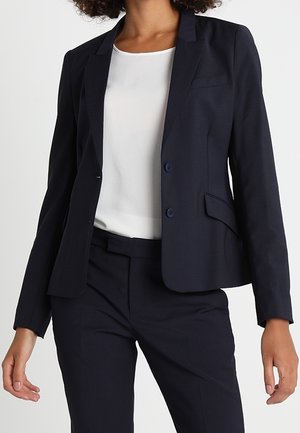 Blazer ajusté bleu marine avec un col cranté, deux boutons et des poches latérales, porté sur un chemisier blanc et assorti à un pantalon.