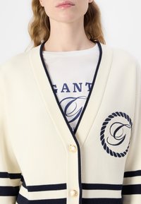 Cardigan bej cu dungi bleumarin, design cu decolteu în V și nasturi aurii. Prezintă un patch cu logo și detalii din frânghie pe partea stângă a pieptului.