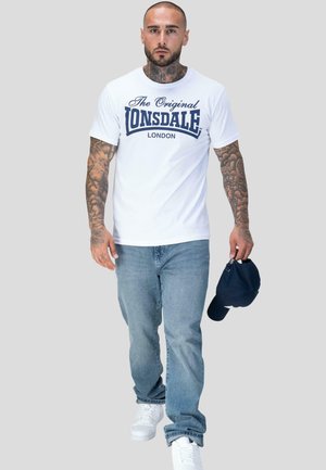 Hombre con tatuajes que lleva una camiseta blanca de Lonsdale London, vaqueros azules y zapatillas blancas, sosteniendo una gorra azul marino en su mano derecha.