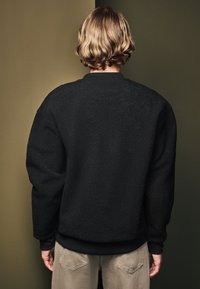 Zwarte fleece sweatshirt met een textuurafwerking, ribgebreide manchetten en onderrand. Draagt beige broek en wordt van de achterkant getoond tegen een donkergroene achtergrond.