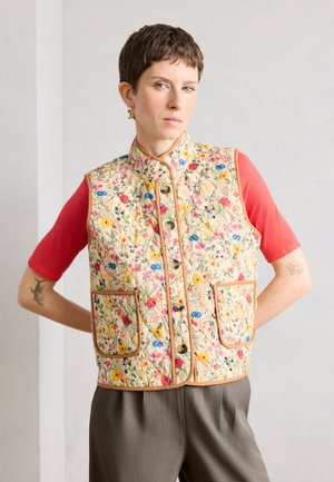 Kortklippet person iført en blomsterquiltet vest med knapper over en koralfarvet kortærmet skjorte, står med hænderne på hoften.