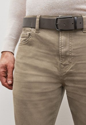 Pantalons en coton beige avec une texture lisse, dotés de poches latérales et d'une ceinture en simili cuir grise avec une boucle noire rectangulaire.