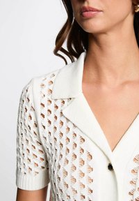 Blusa bianca a maniche corte in maglia con un dettagliato motivo a rete, colletto e chiusura con un solo bottone. Tessuto testurizzato in tutto.