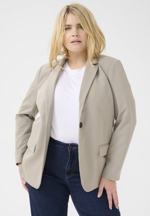 Beige sako s jedním knoflíkem, klopami a dvěma předními kapsami, nošené přes bílé tričko a tmavé denimové džíny. Měkká textura.