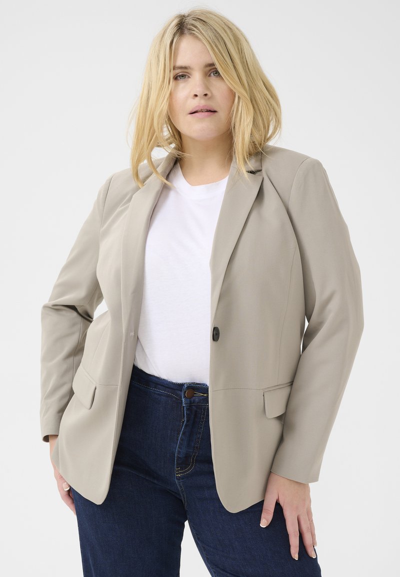 Blazer beige con un solo bottone, a bavero, e due tasche anteriori, indossato sopra una camicia bianca e jeans in denim scuro. Tessuto morbido.