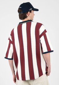 Jeune homme portant une casquette bleu marine et un pull en maille à manches courtes avec des rayures verticales bordeaux et blanches, vu de dos.