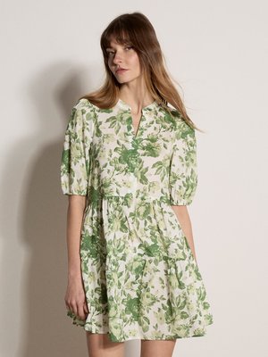 Vrouw draagt een korte, losse groene en witte jurk met bloemenprint, pofmouwen en een kleine V-hals aan de voorkant tegen een effen achtergrond.