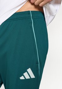 Tirkio spalvos sportinės šortai iš lygaus audinio, su šonine kišene, kontrastinga šviesiai mėlyna juostele ir baltu Adidas logotipu ant krašto.