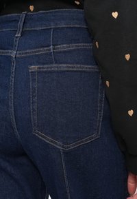 Tumedatest sinised denim püksid, millel on klassikaline disain ning tagakotil on kontrastne õmblemise joon. Tekstuur näib sujuv ja vastupidav.
