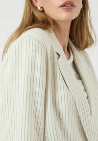 Lichtgekleurde blazer met verticale streepjes, gestructureerde schouders en een notch revers. De stof heeft een zachte textuur met een subtiele glans.