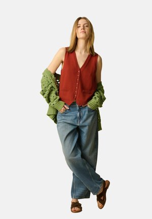Femme debout, les mains dans les poches, portant un gilet rouille boutonné, un cardigan vert en grosse maille qui glisse des épaules, un jean ample et des sandales marron.