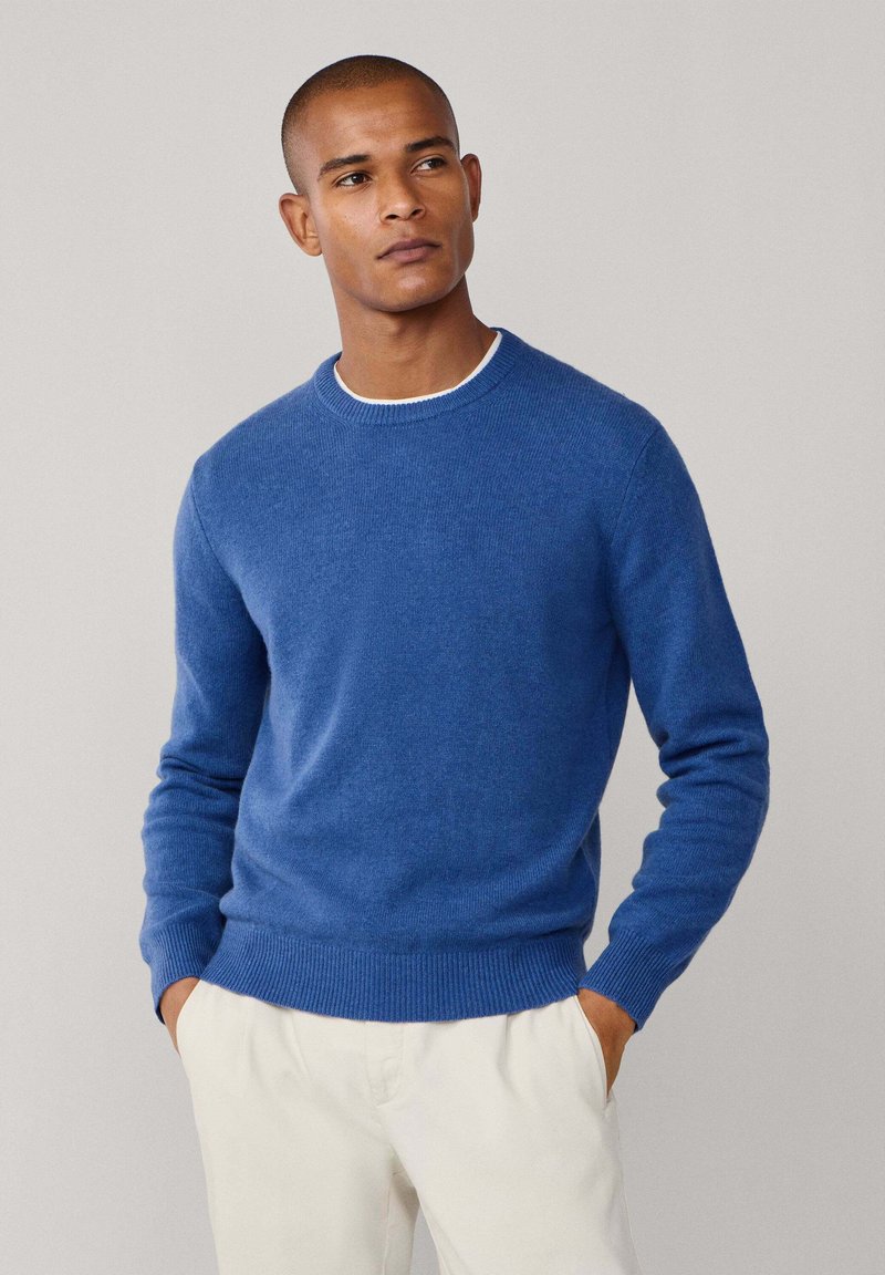 Blå, rundhalsad tröja i mjukt, texturerat stickat material. Har ribbade ärmslut och midja, ihopparad med ljusa byxor.